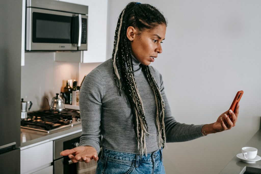 A woman standing in her kitchen, looking confused and frustrated while holding a phone, symbolizing gaslighting as on of the signs of a narcissistic mother.