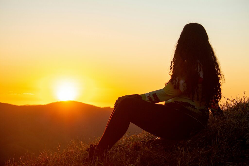 A woman sitting on a hill at sunset, reflecting on her thoughts in a  peaceful setting as her healing journey begins.