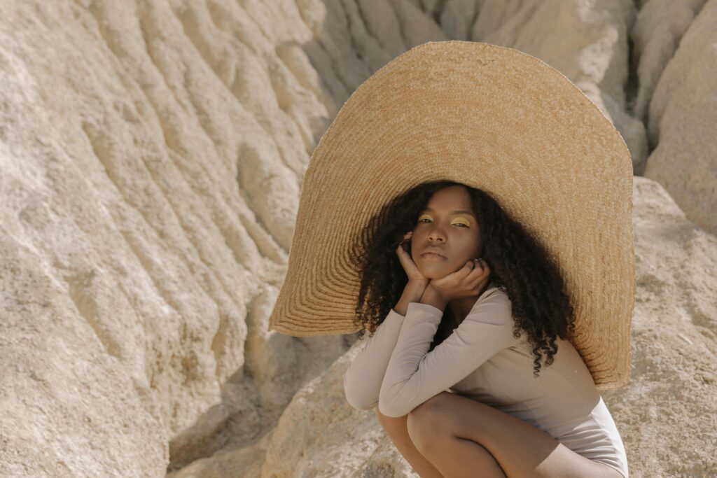 A woman crouching in a desert landscape, wearing a large straw hat, symbolizing protection and introspection.