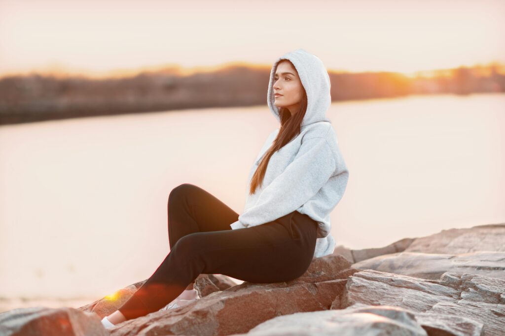 A woman in a gray hoodie sitting on rocks near water, appearing calm and reflective after realizing narcissistic abuse.